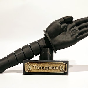 May include: A black 3D printed sculpture of a hand with a segmented arm attached to a base with a gold plaque that reads "Thompson".