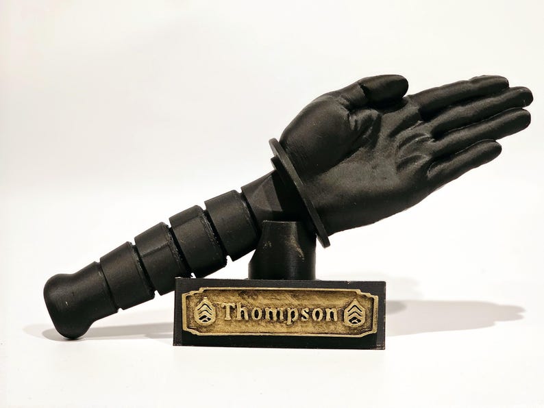 May include: A black 3D printed sculpture of a hand with a segmented arm attached to a base with a gold plaque that reads "Thompson".
