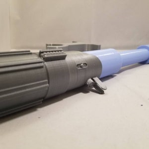 May include: A 3D-printed, futuristic-looking device with a cylindrical design. The device features a gray, ridged section, a gray mid-section with a lever, and a light blue, elongated tube with a rounded end. The object is likely a prop or model.