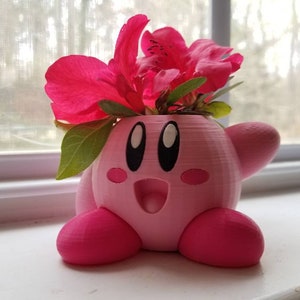 May include: A pink 3D printed planter shaped like the character Kirby from the video game series Kirby. The planter has a flower arrangement of pink azalea flowers.