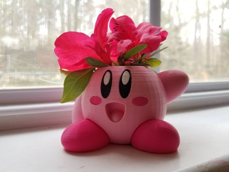 May include: A pink 3D printed planter shaped like the character Kirby from the video game series Kirby. The planter has a flower arrangement of pink azalea flowers.