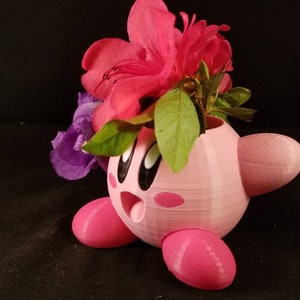 May include: A pink 3D printed planter shaped like a cartoon character with flowers in it. The planter has a round body with two small arms and legs. The flowers are pink and purple.
