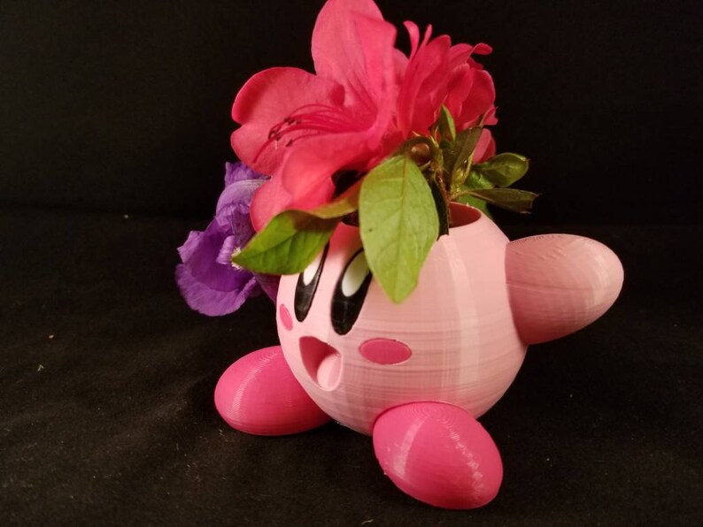 May include: A pink 3D printed planter shaped like a cartoon character with flowers in it. The planter has a round body with two small arms and legs. The flowers are pink and purple.