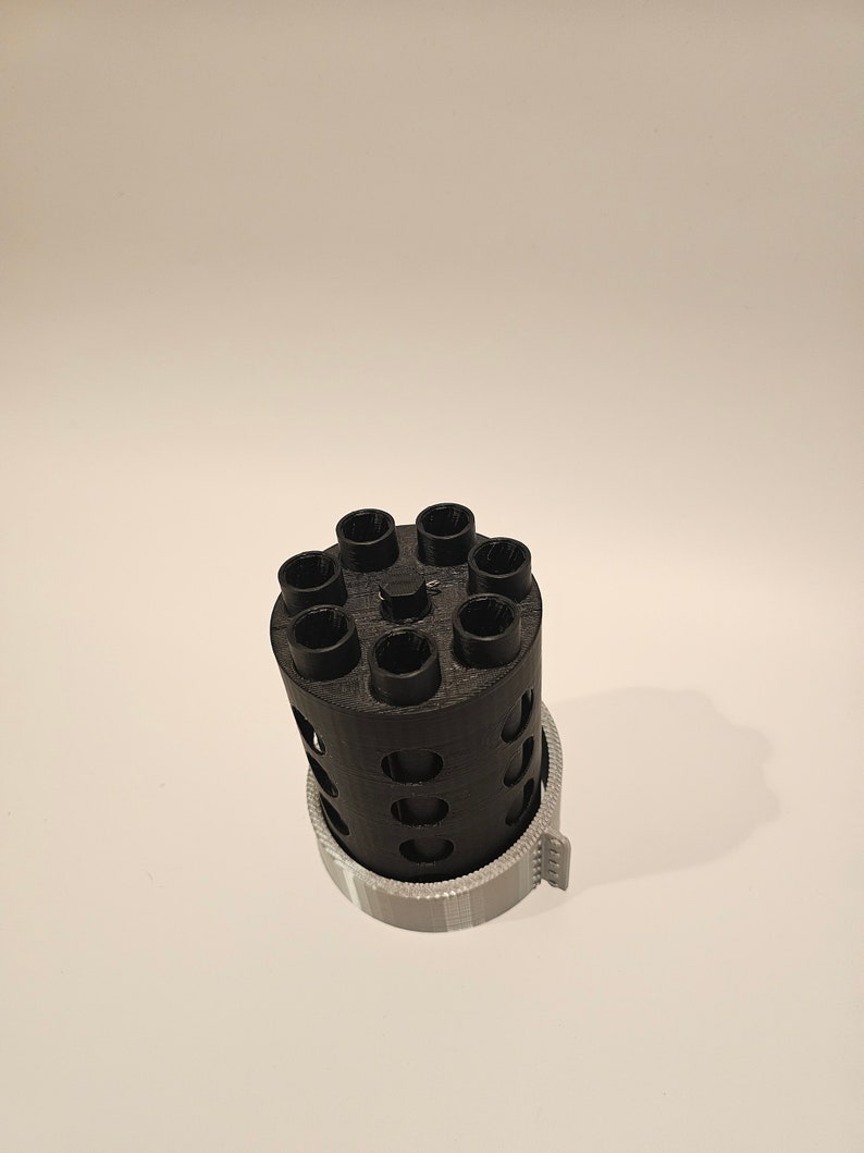 May include: A black 3D printed cylinder with eight holes around the top and a gray plastic ring around the bottom.