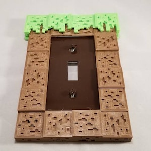 May include: A light switch cover with a Minecraft-themed design. The cover is made of brown plastic and features a green grass block on top. The switch is visible in the center of the cover.