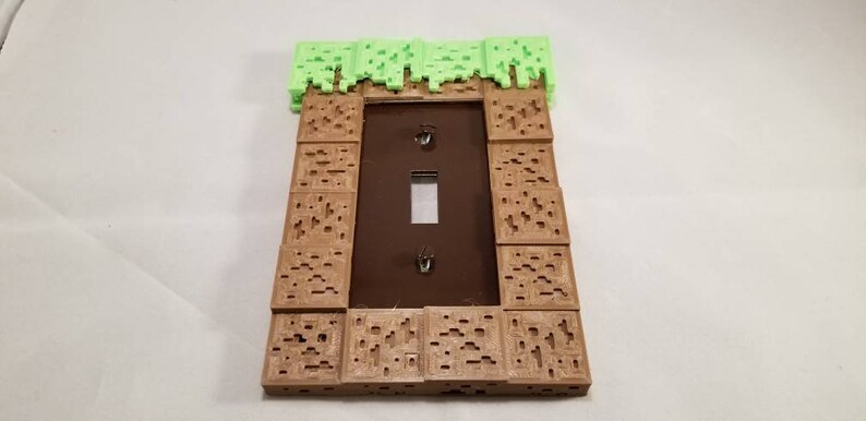 May include: A light switch cover with a Minecraft-themed design. The cover is made of brown plastic and features a green grass block on top. The switch is visible in the center of the cover.