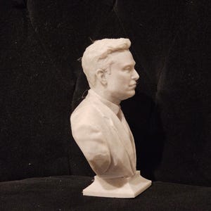 May include: A white bust sculpture of a person wearing a suit jacket. The sculpture is on a white base.