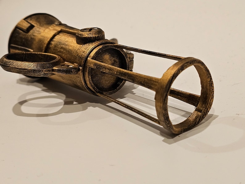 May include: A gold-colored 3D printed model of a futuristic weapon with a cylindrical body and a cage-like structure at the end.