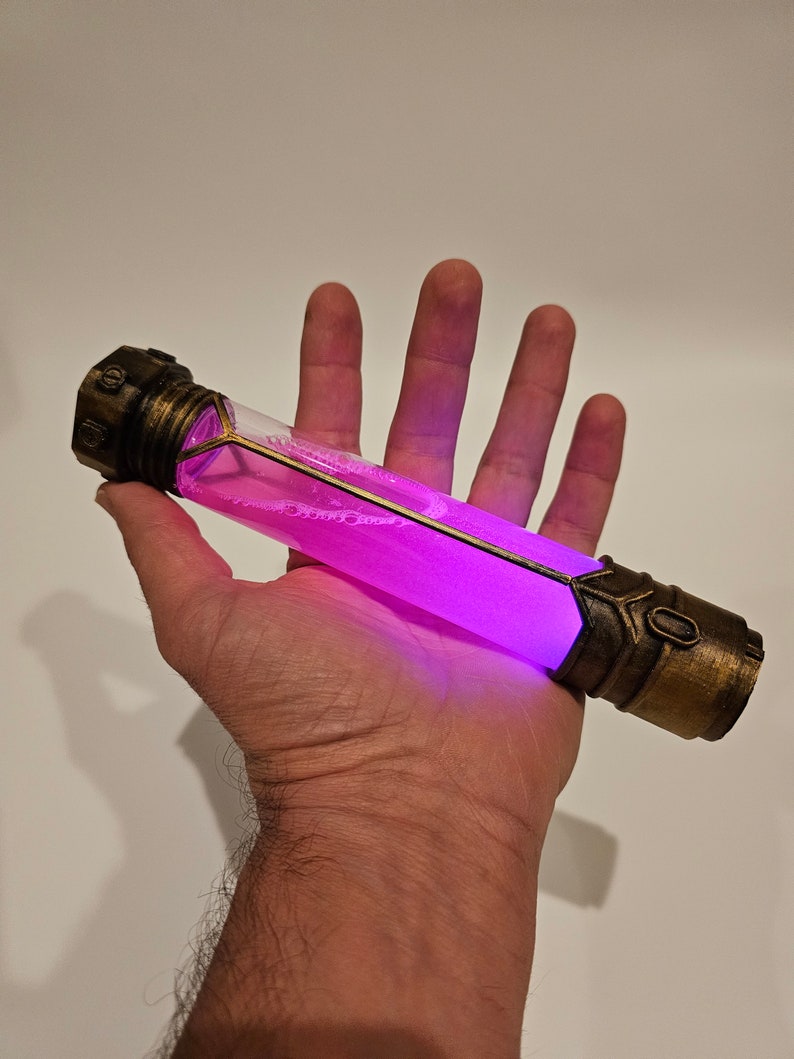 May include: A purple glowing liquid inside a clear glass tube with gold metal accents. The tube is held in a hand.