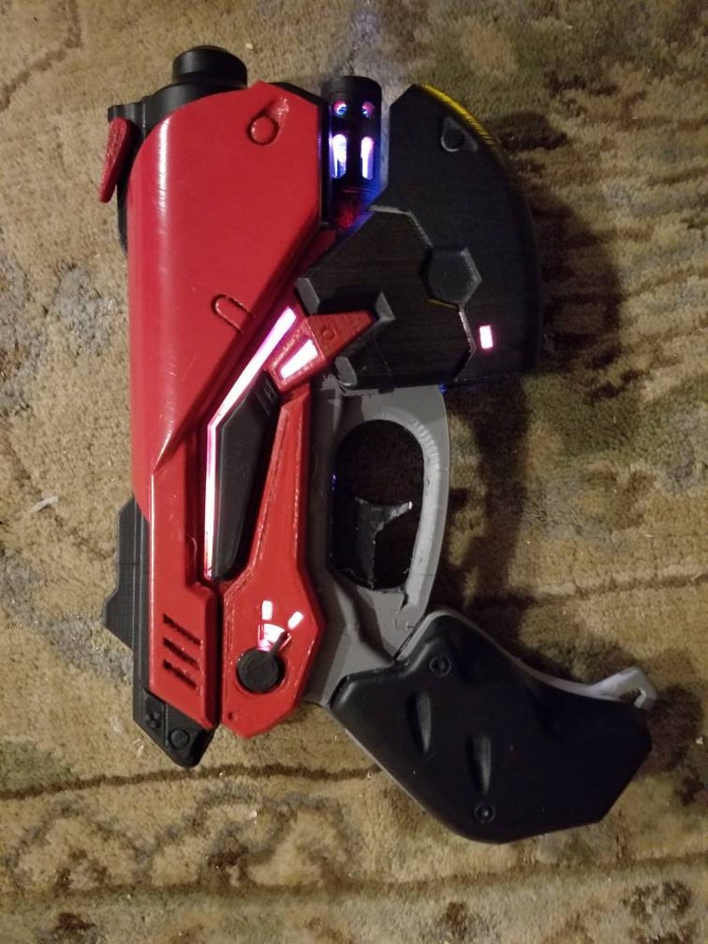 Puede incluir: Una pistola de juguete de pl&aacute;stico roja y negra con un gatillo blanco y un mango negro. La pistola tiene un dise&ntilde;o futurista con luces rojas y azules brillantes.