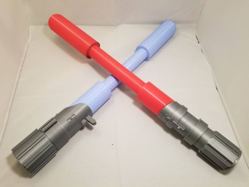May include: Two toy lightsabers crossed. One is red with a gray handle, the other is light blue with a gray handle. The lightsabers are made of plastic and are designed to look like the iconic weapons from a science fiction franchise.