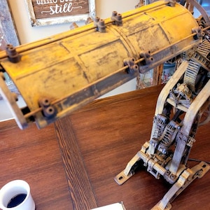 Puede incluir: Una l&aacute;mpara de estilo steampunk con una pantalla de metal amarilla y engranajes y palancas intrincados. La l&aacute;mpara est&aacute; hecha de metal y tiene un aspecto r&uacute;stico e industrial.