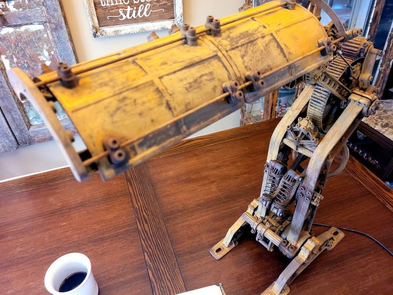 Puede incluir: Una l&aacute;mpara de estilo steampunk con una pantalla de metal amarilla y engranajes y palancas intrincados. La l&aacute;mpara est&aacute; hecha de metal y tiene un aspecto r&uacute;stico e industrial.