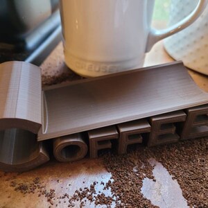 May include: A brown 3D printed coffee holder with the word "COFFEE" spelled out in raised letters. The holder is designed to hold a coffee mug or cup.