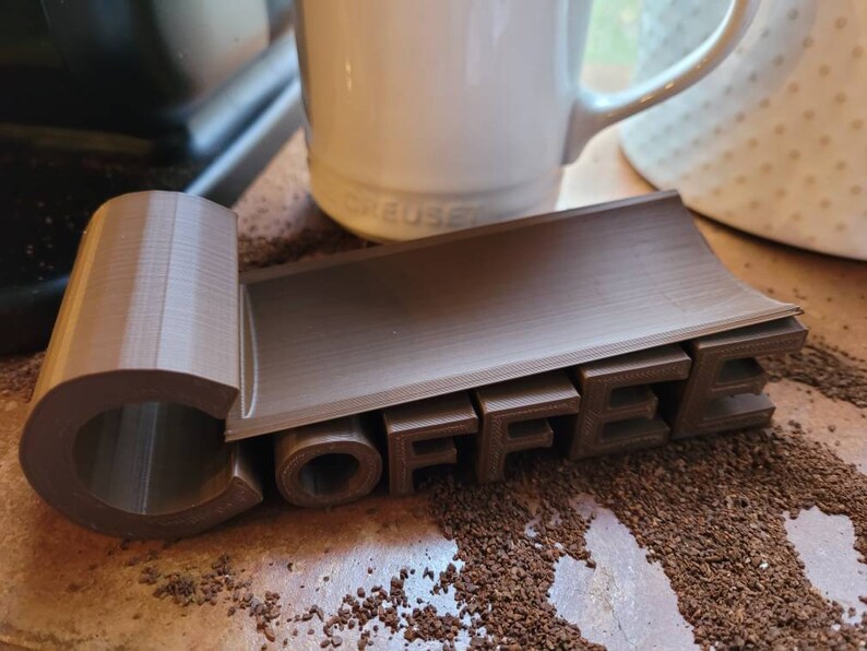 May include: A brown 3D printed coffee holder with the word "COFFEE" spelled out in raised letters. The holder is designed to hold a coffee mug or cup.