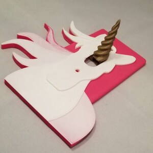 May include: A white unicorn light switch cover with a gold spiral horn. The cover is mounted on a pink background.