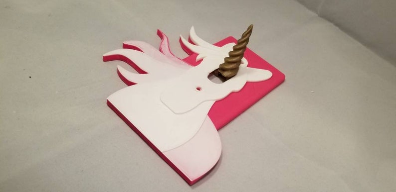 May include: A white unicorn light switch cover with a gold spiral horn. The cover is mounted on a pink background.