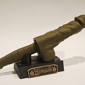 May include: An olive-green sculpture of a phallus, mounted on a black rectangular base. The base features a gold-colored nameplate with the name "Thompson" and military insignia. The sculpture is detailed and textured.