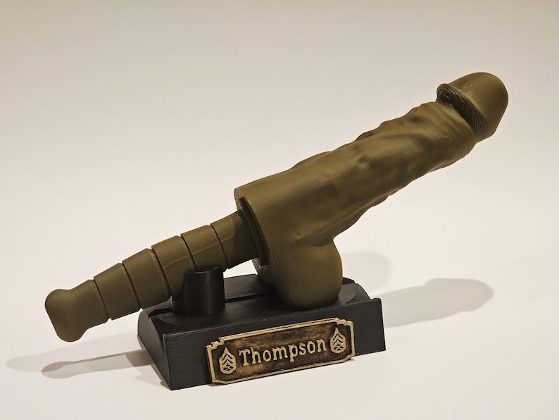 May include: An olive-green sculpture of a phallus, mounted on a black rectangular base. The base features a gold-colored nameplate with the name "Thompson" and military insignia. The sculpture is detailed and textured.