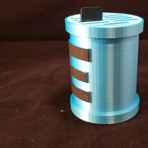 May include: A light blue 3D printed storage container with a black grid on top and black bands around the side. The container is designed to hold small items like SD cards.