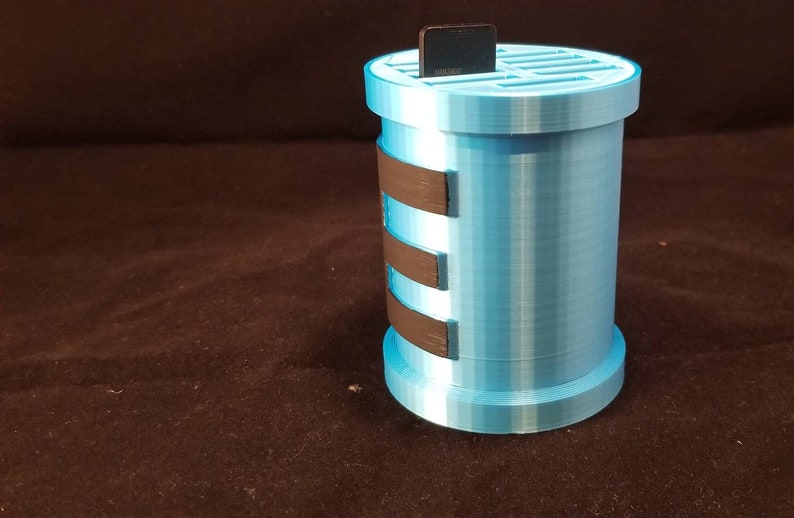 May include: A light blue 3D printed storage container with a black grid on top and black bands around the side. The container is designed to hold small items like SD cards.