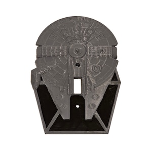 May include: A gray plastic light switch cover shaped like the Millennium Falcon spaceship from Star Wars. The cover has a black background and a detailed design of the spaceship.