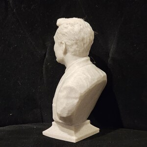 May include: White plaster bust sculpture of a person with short hair. The bust is on a white base.
