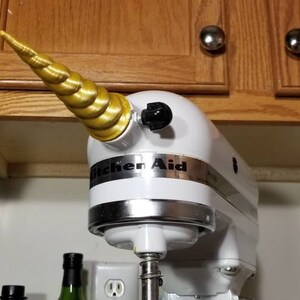 May include: A white KitchenAid stand mixer with a gold unicorn horn attached to the top. The horn is made of a spiral design.