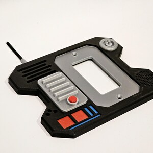 May include: A black and silver 3D printed device with a rectangular screen, a red button, and a silver button panel. The device has a black antenna and a small, round, gray knob on the top.