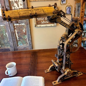 Puede incluir: Una l&aacute;mpara de estilo steampunk con un brazo largo y articulado y una pantalla de metal amarilla. La l&aacute;mpara est&aacute; hecha de engranajes de metal y otras piezas de aspecto industrial. La l&aacute;mpara est&aacute; sentada sobre una mesa de madera con un libro y una taza de caf&eacute;.