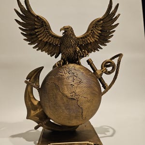 May include: A bronze-colored sculpture featuring an eagle with outstretched wings atop a globe, an anchor, and a rope. The base has the name "Smith" and military insignia. The sculpture is a decorative item.