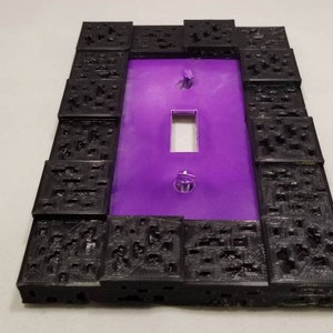 May include: A decorative light switch cover with a purple center and a black, textured border. The cover has a rectangular opening for the switch and two screws for mounting. The border has a unique, geometric design.
