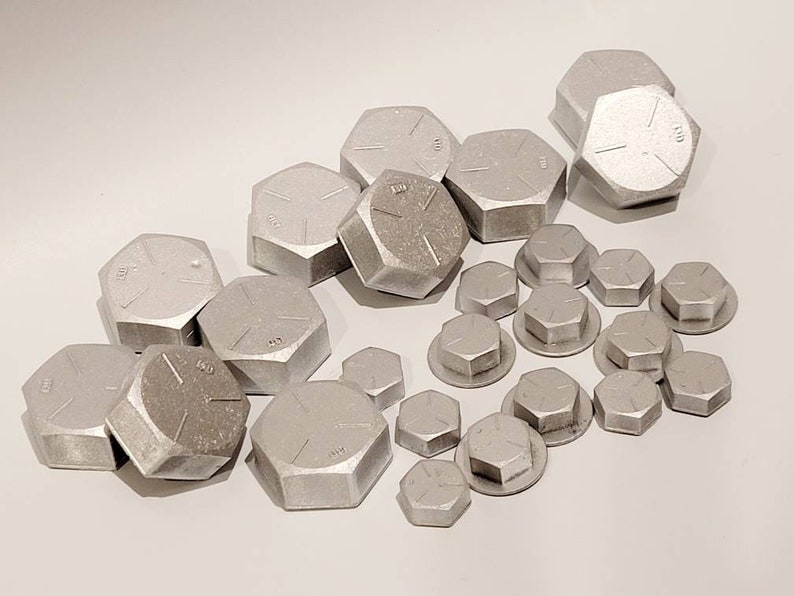 K&ouml;nnte beinhalten: Eine Ansammlung silberfarbener, sechseckiger Metallkappen in verschiedenen Gr&ouml;&szlig;en. Einige Kappen weisen Markierungen auf ihren flachen Oberfl&auml;chen auf. Die Kappen sind auf einer hellen Oberfl&auml;che verstreut, was auf ein Industrie- oder Hardware-Thema hindeutet.