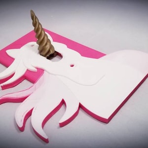 May include: A pink and white unicorn light switch cover with a gold horn. The unicorn is outlined in pink and the horn is a 3D element.