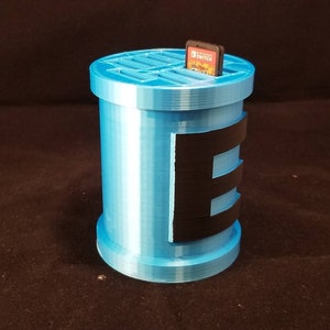 May include: A light blue 3D printed game cartridge holder with a black stripe design and a slot for a Nintendo Switch game cartridge. The holder is designed to hold multiple game cartridges.