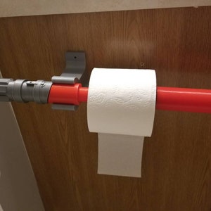 May include: A novelty toilet paper holder designed to resemble a lightsaber. The holder features a gray and red design, with a roll of white toilet paper in place. The holder is mounted on a wooden surface.