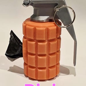 May include: A pink grenade with a gray top and pull ring. The body of the grenade is a textured, rectangular design. A black object is attached to the side. The word "Pink" is written in pink at the bottom.