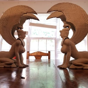 May include: Two beige ceramic sculptures of winged women with human heads and lion bodies. The sculptures are facing each other and have their wings spread out.