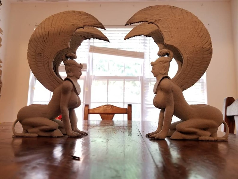 May include: Two beige ceramic sculptures of winged women with human heads and lion bodies. The sculptures are facing each other and have their wings spread out.