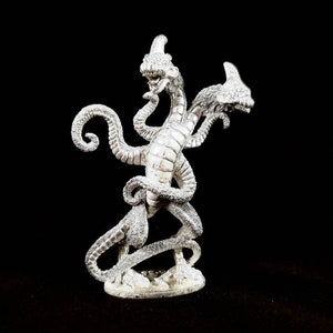Op de afbeelding: Een zilverkleurig, driekoppig drakenbeeldje met een gedetailleerd, gestructureerd oppervlak. De draak heeft twee koppen die naar voren gericht zijn en één kop die naar achteren gericht is. Het beeldje staat op een ronde basis.
