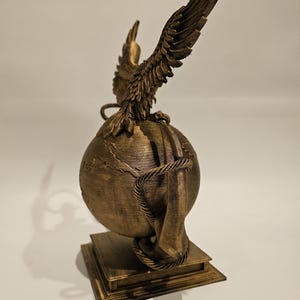 May include: A bronze-colored sculpture featuring an eagle with outstretched wings perched atop a globe. The globe is encircled by a rope and rests on a square base. The sculpture is detailed and appears to be made of a metallic material.