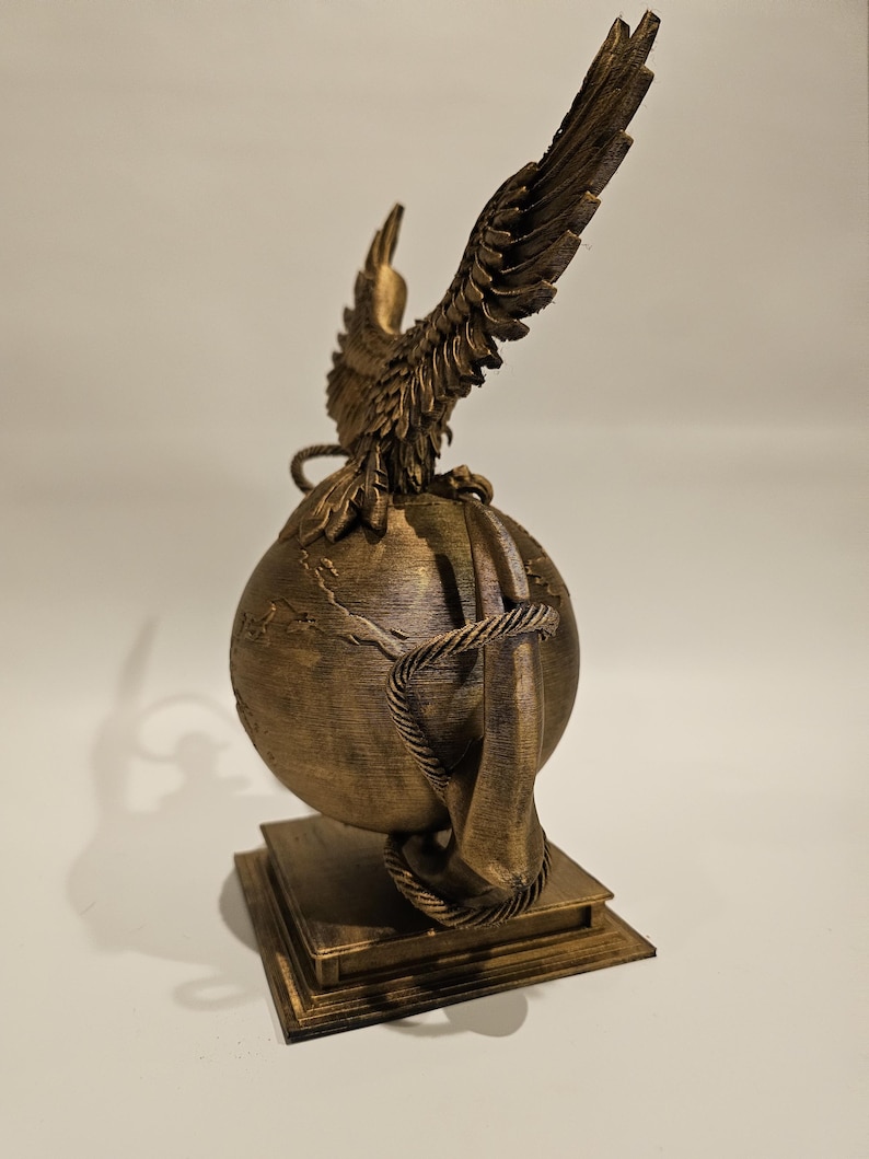 May include: A bronze-colored sculpture featuring an eagle with outstretched wings perched atop a globe. The globe is encircled by a rope and rests on a square base. The sculpture is detailed and appears to be made of a metallic material.