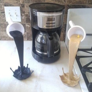 May include: A black coffee maker with a white ceramic coffee pot pouring black coffee into a white ceramic cup. A second white ceramic coffee pot is pouring light brown coffee into a white ceramic cup.