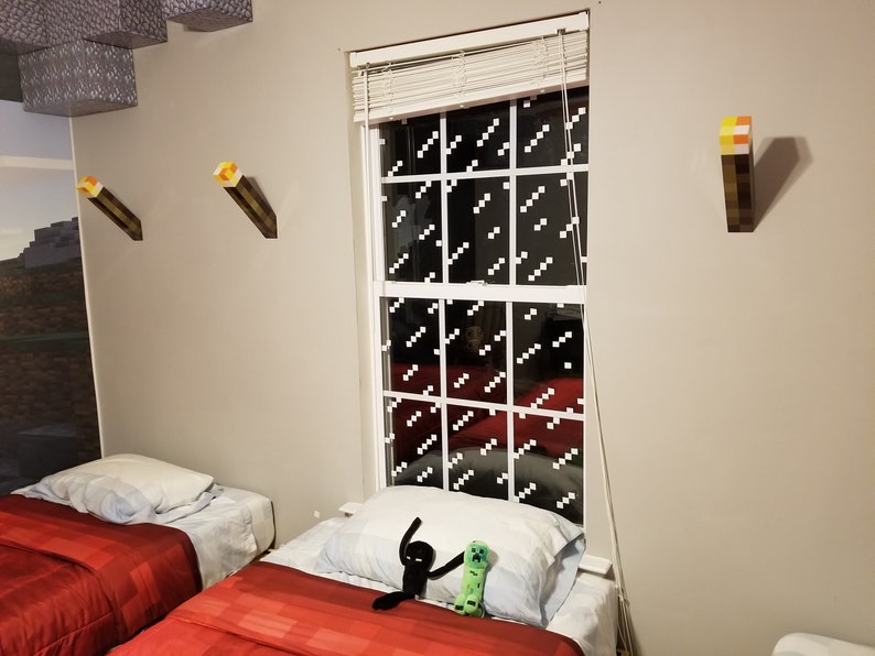 May include: A bedroom with two twin beds, red bedding, and a window with a Minecraft-themed design. There are three Minecraft torch wall decorations, two black and green Minecraft character toys on the bed, and a Minecraft-themed mural on the wall.