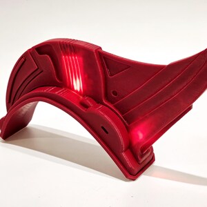 Op de afbeelding: Een rood plastic accessoire met een gebogen vorm en een licht dat een rode gloed uitstraalt. Het accessoire heeft een geometrisch ontwerp en lijkt deel uit te maken van een kostuum of cosplay.