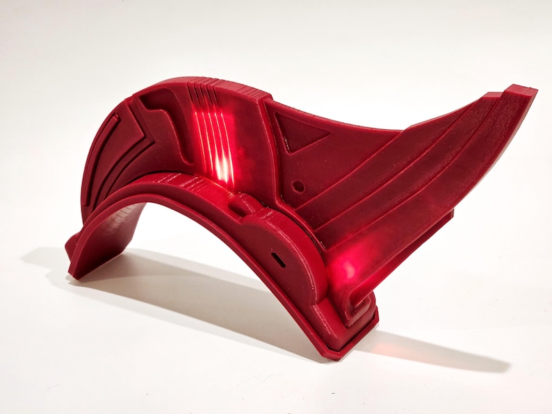 Op de afbeelding: Een rood plastic accessoire met een gebogen vorm en een licht dat een rode gloed uitstraalt. Het accessoire heeft een geometrisch ontwerp en lijkt deel uit te maken van een kostuum of cosplay.