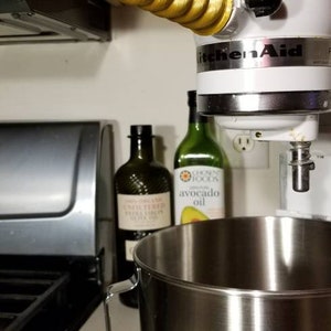 May include: A white KitchenAid stand mixer with a gold unicorn horn attached to the top. The mixer has a stainless steel bowl and is sitting on a white counter. There are two bottles of oil behind the mixer.