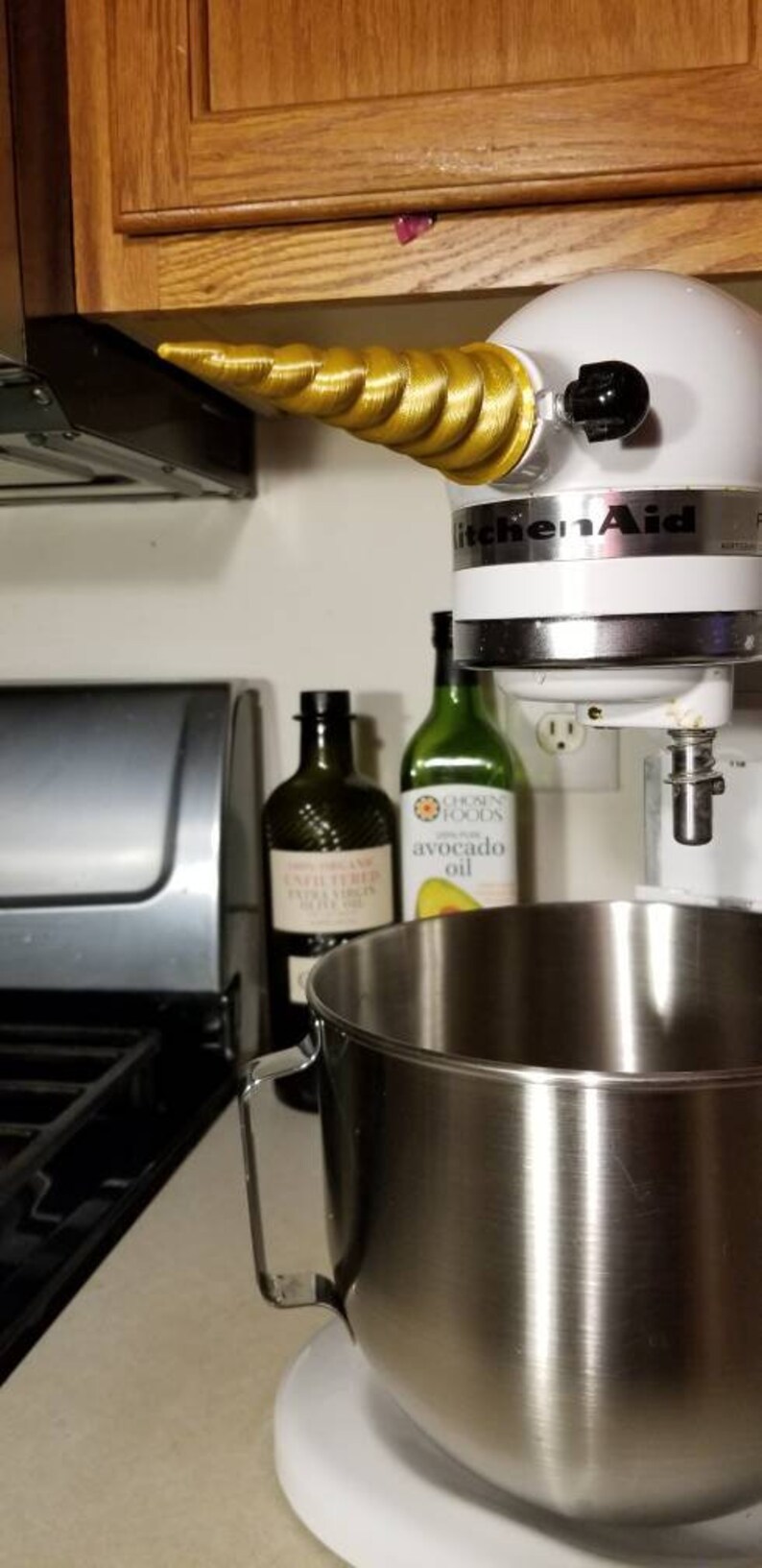 May include: A white KitchenAid stand mixer with a gold unicorn horn attached to the top. The mixer has a stainless steel bowl and is sitting on a white counter. There are two bottles of oil behind the mixer.