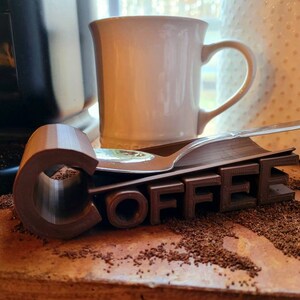 May include: A brown 3D printed word "COFFEE" with a spoon resting on top. The word is sitting on a brown surface with coffee grounds scattered around it.