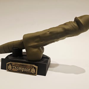 May include: A khaki-colored sculpture of a phallus mounted on a black base. The base features a gold-colored nameplate with the name "Thompson" and military insignia. The sculpture is detailed and realistic.